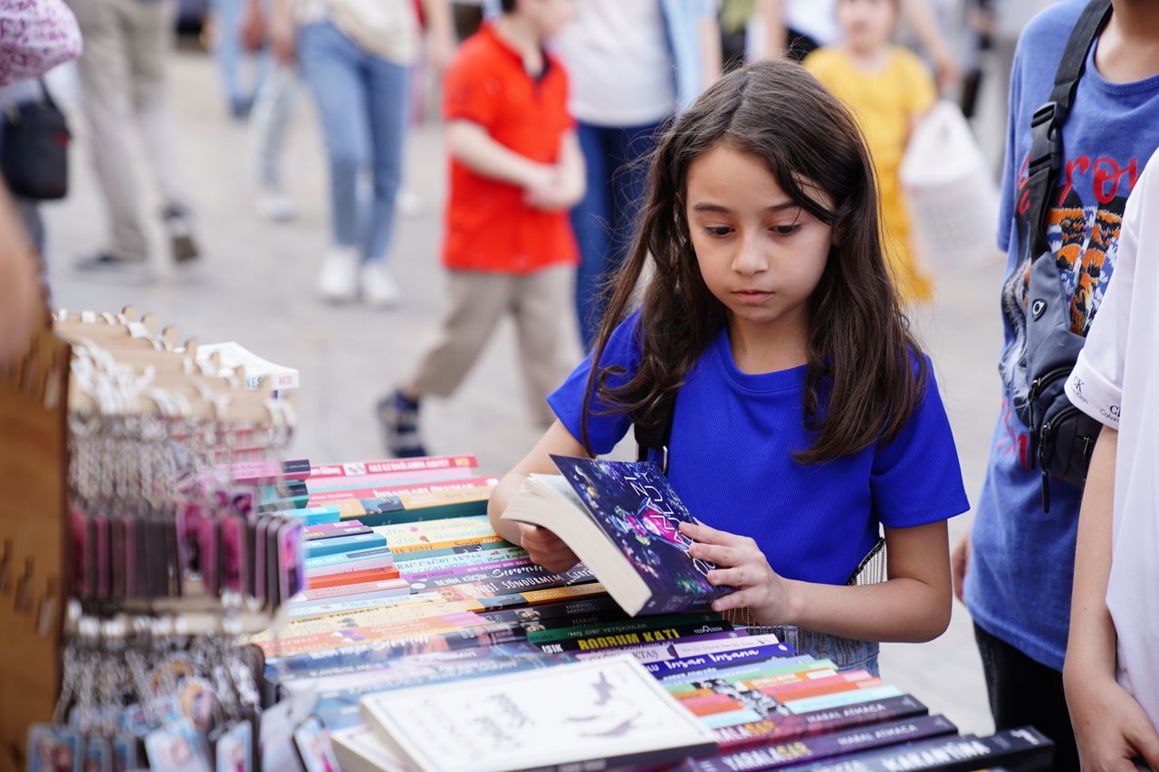Young girl reading at a colorful outdoor book market, exploring books.